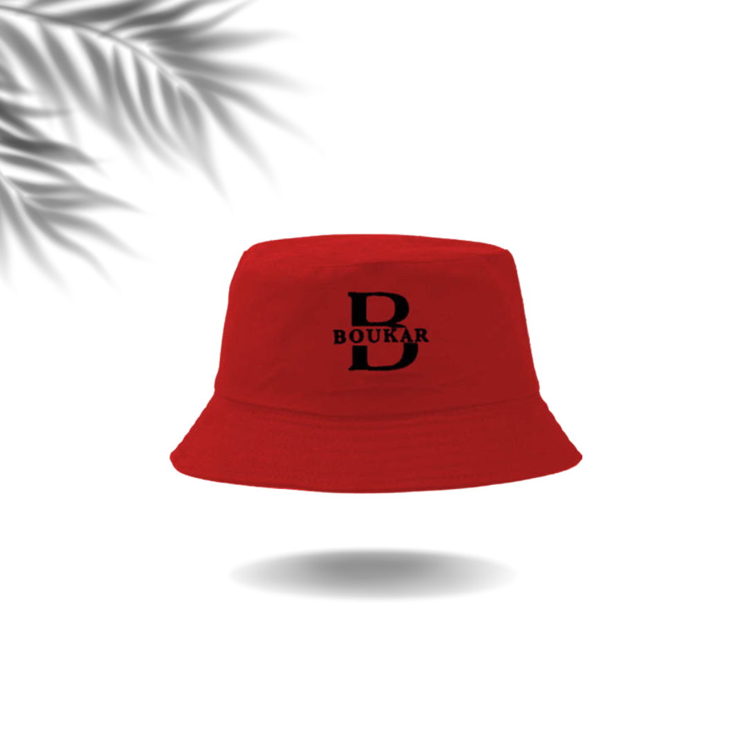 Red bucket hat with black embroidered text 'D BOUKAR' on the front, floating above a gray shadow on a white background, with a partial gray palm leaf in the top left corner.