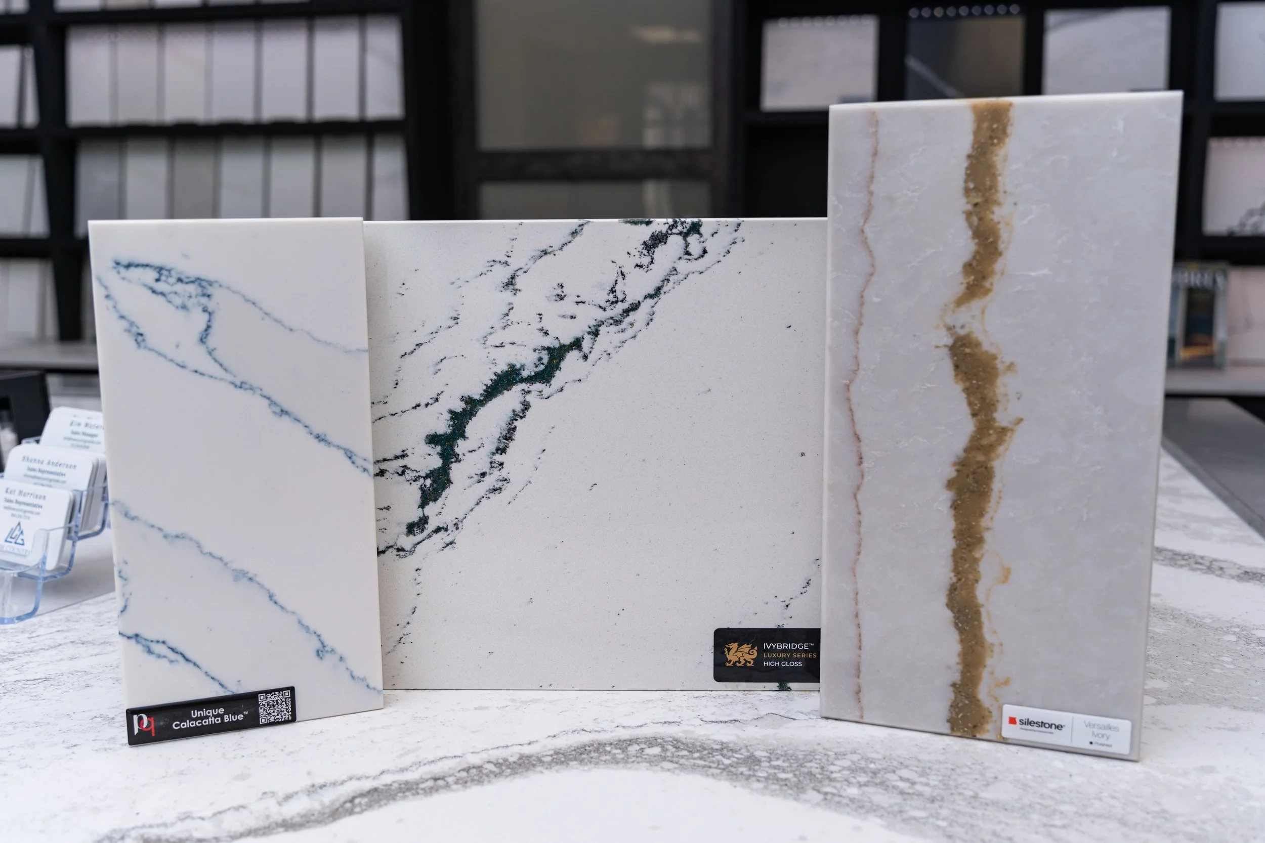 Three upright showroom slabs: white with blue veining ("Unique Calacatta Blue"), white with dark green veining ("Ivybridge Luxury Series"), and white with a gold vein ("Silestone Versailles Ivory").