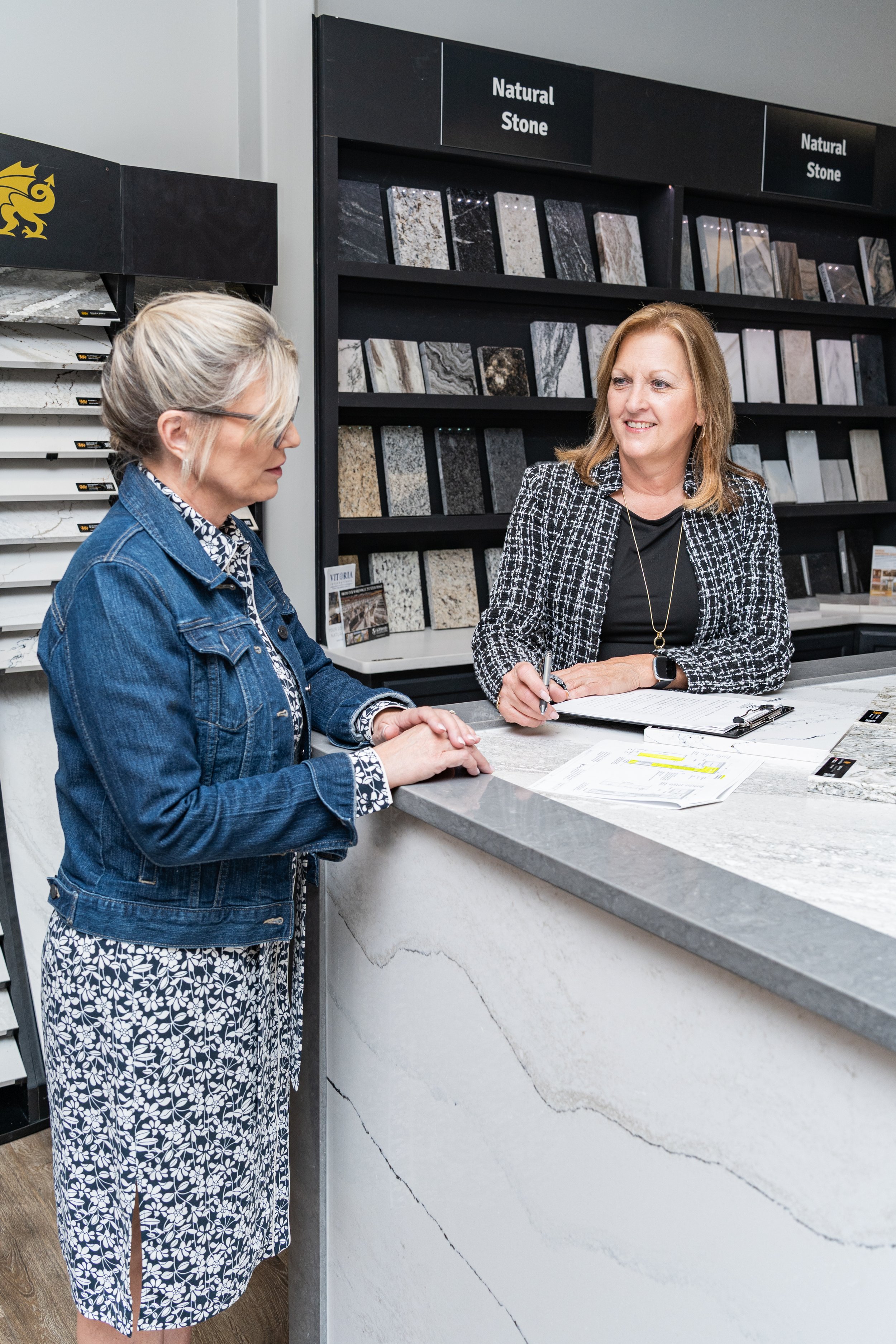Two women speaking at a showroom counter, with a sales representative writing on paperwork while a customer looks on, in front of shelves of "Natural Stone" sample tiles.