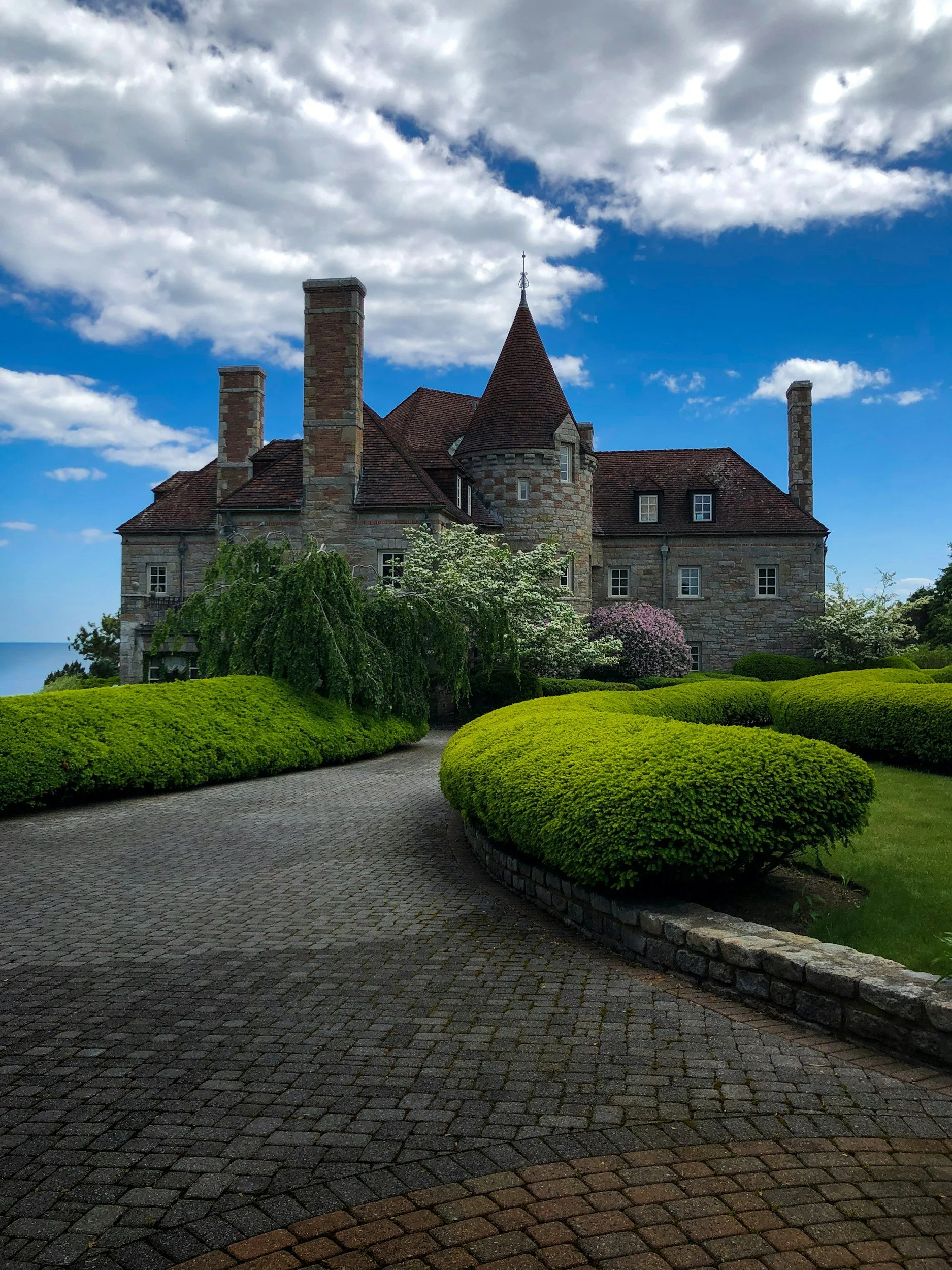 A stone castle with turret and chimneys, surrounded by lush green shrubs and trees, under a partly cloudy blue sky.