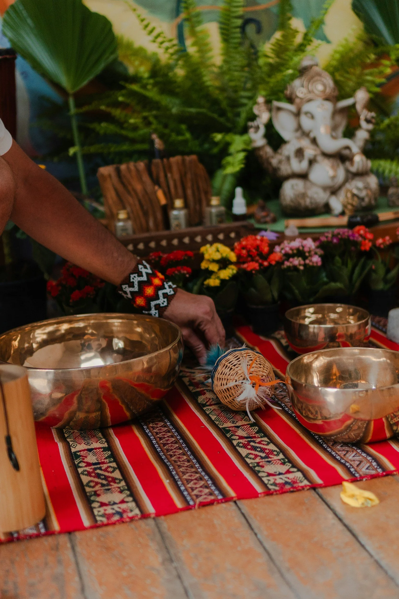 A close-up photo of a colorful striped tablecloth with metallic singing bowls, a small woven basket with feathers, a few yellow flower petals, and some other objects in a setting with green plants and a large decorative stone statue of Ganesha in the background.
