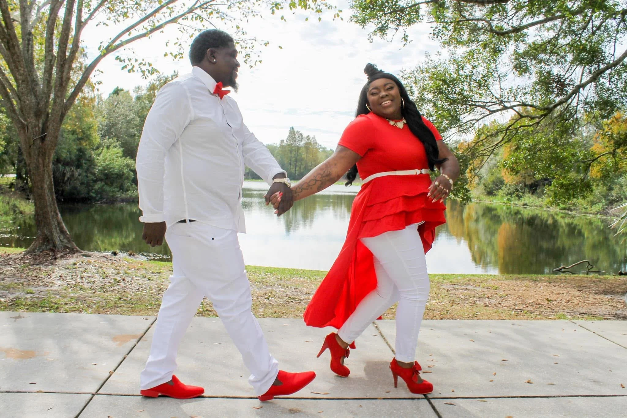 A couple holding hands and dancing outdoors by a lake, with the woman wearing a red dress and the man in white attire with red shoes and bowtie.