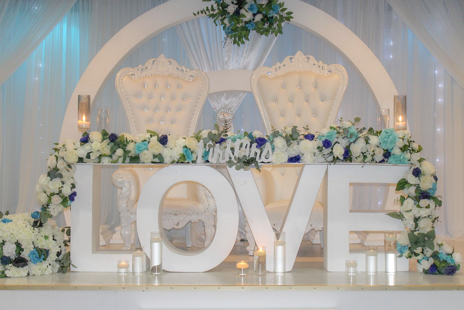 Wedding stage with two ornate white chairs, surrounded by white and blue flowers, candles, and a decorative sign reading 'LOVE'.