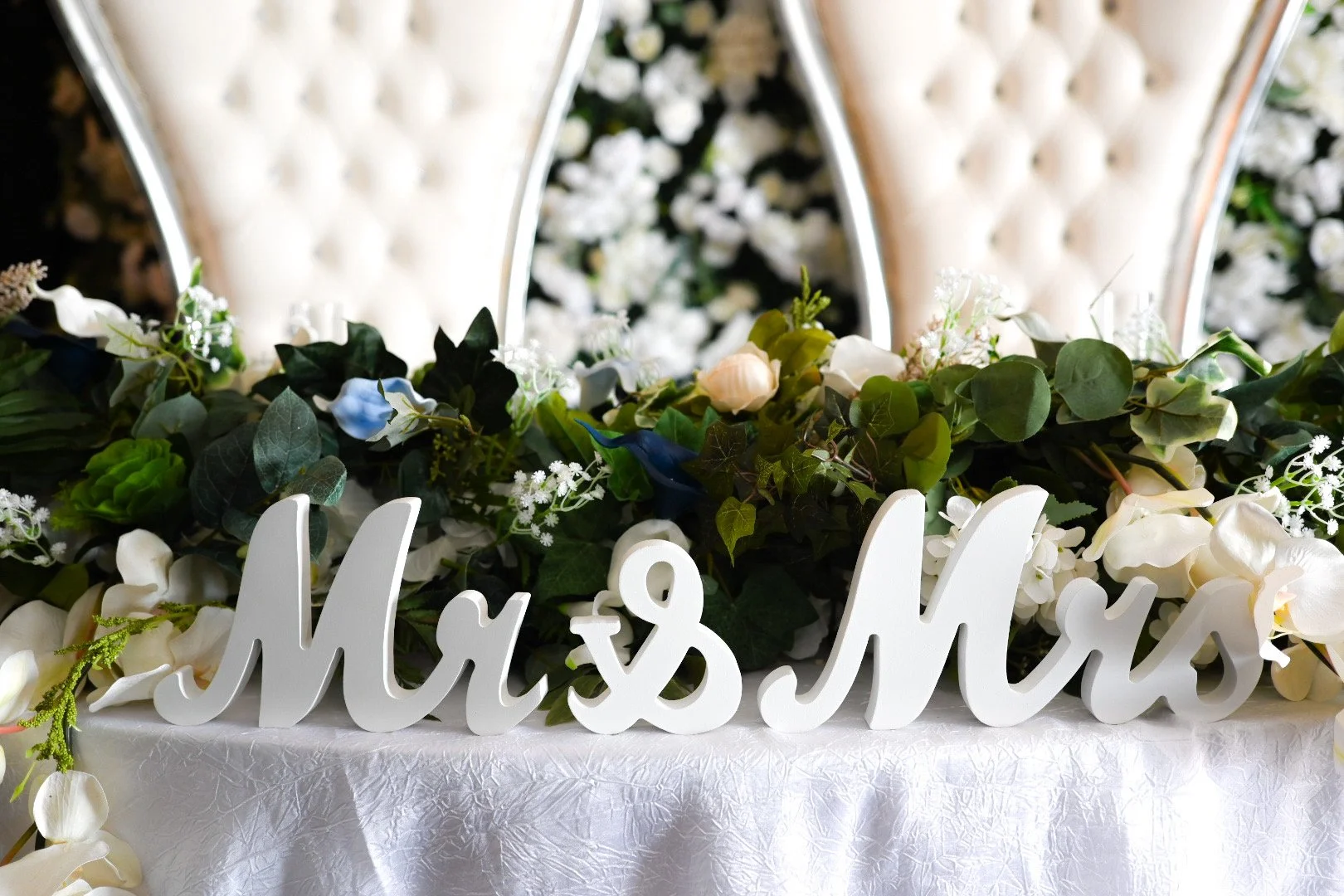 White decorative sign reading 'Mrs & Mr' on a floral arrangement with white and green flowers and greenery, set on a white tablecloth with ornate high-backed white chairs in the background.