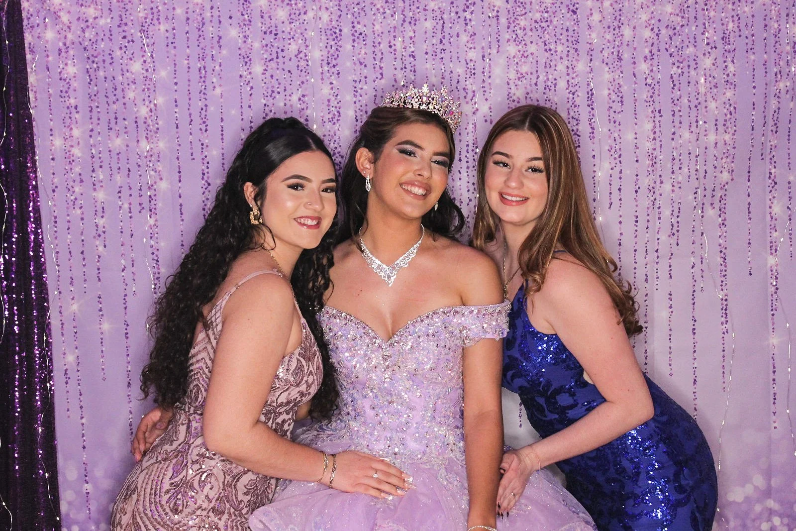 Three young women dressed in glamorous evening gowns at a quinceañera celebration, one wearing a tiara, standing in front of a purple curtain backdrop with hanging purple and silver decorations.