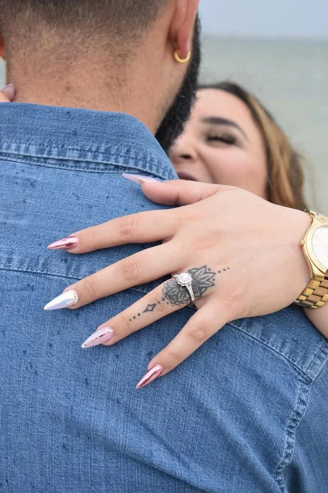 A woman hugging a man, showing her hand with a tattoo, a diamond ring, a gold watch, and long manicured nails, at the beach.