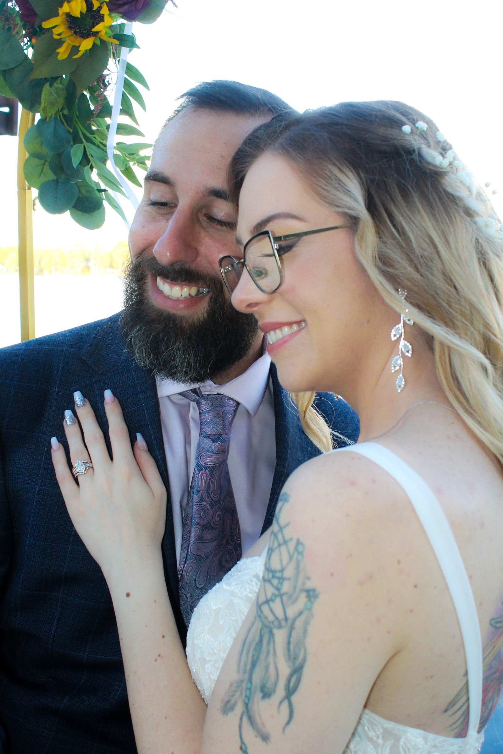 A smiling bride and groom embrace closely outdoors, with the bride wearing glasses, earrings, and a wedding dress, and the groom in a suit and tie, surrounded by green foliage and flowers.