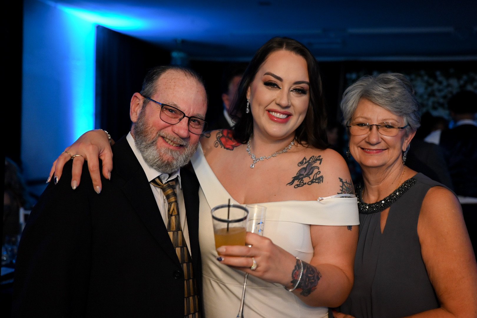 Three people smiling and posing together at a celebration or event. The woman in the middle is holding a drink, has dark hair, tattoos, and is dressed in a white off-shoulder dress. The man on the left has a beard, glasses, and is wearing a black sui