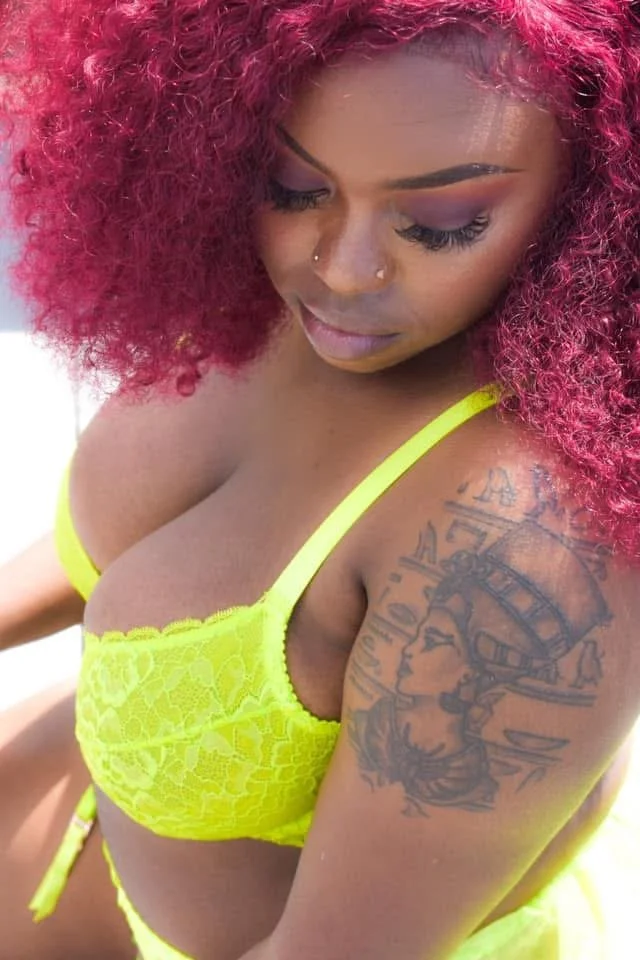 A woman with curly, bright red hair wearing a neon yellow lace bra, showing a tattoo on her upper arm of a woman's profile with a compass and clock in the background.
