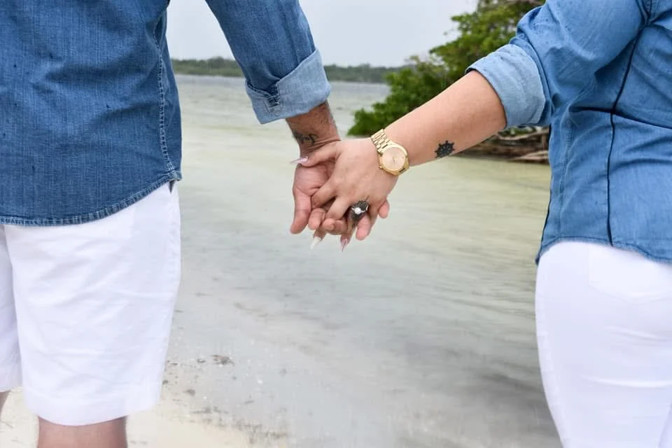 Two people holding hands at a beach with water and trees in the background.