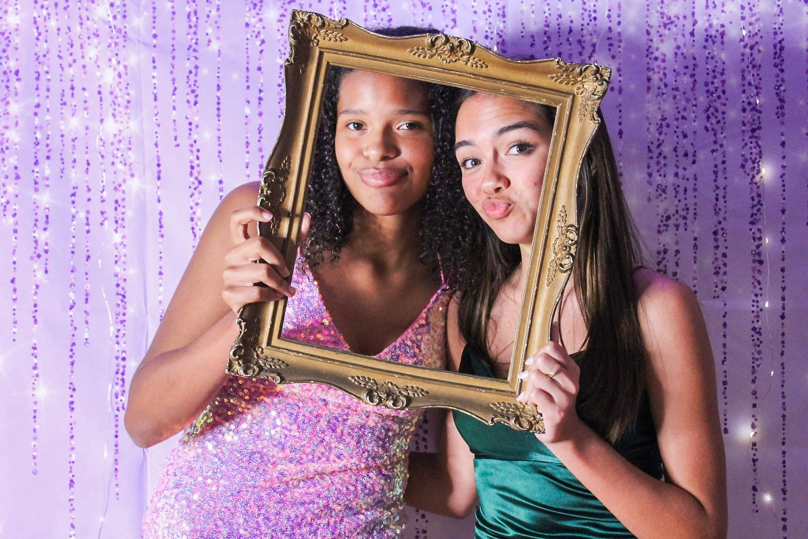 Two young women holding a decorative gold picture frame, standing in front of a purple curtain with hanging beads, one wearing a pink sequined dress and the other in a green satin dress, posing together at a party.