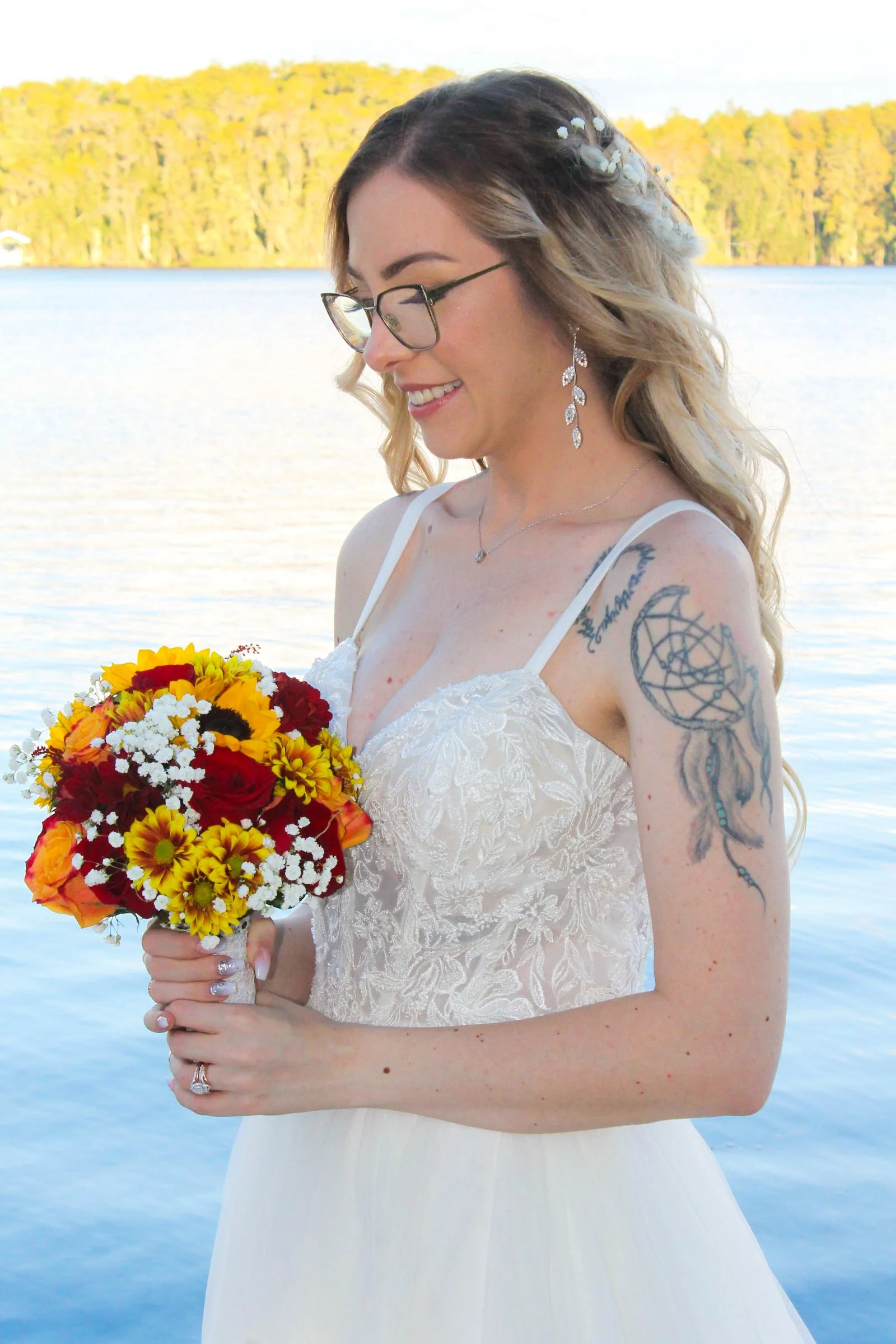 A smiling woman in a white dress holding a bouquet of yellow, red, and orange flowers, standing near a body of water with trees in the background, wearing glasses and jewelry.