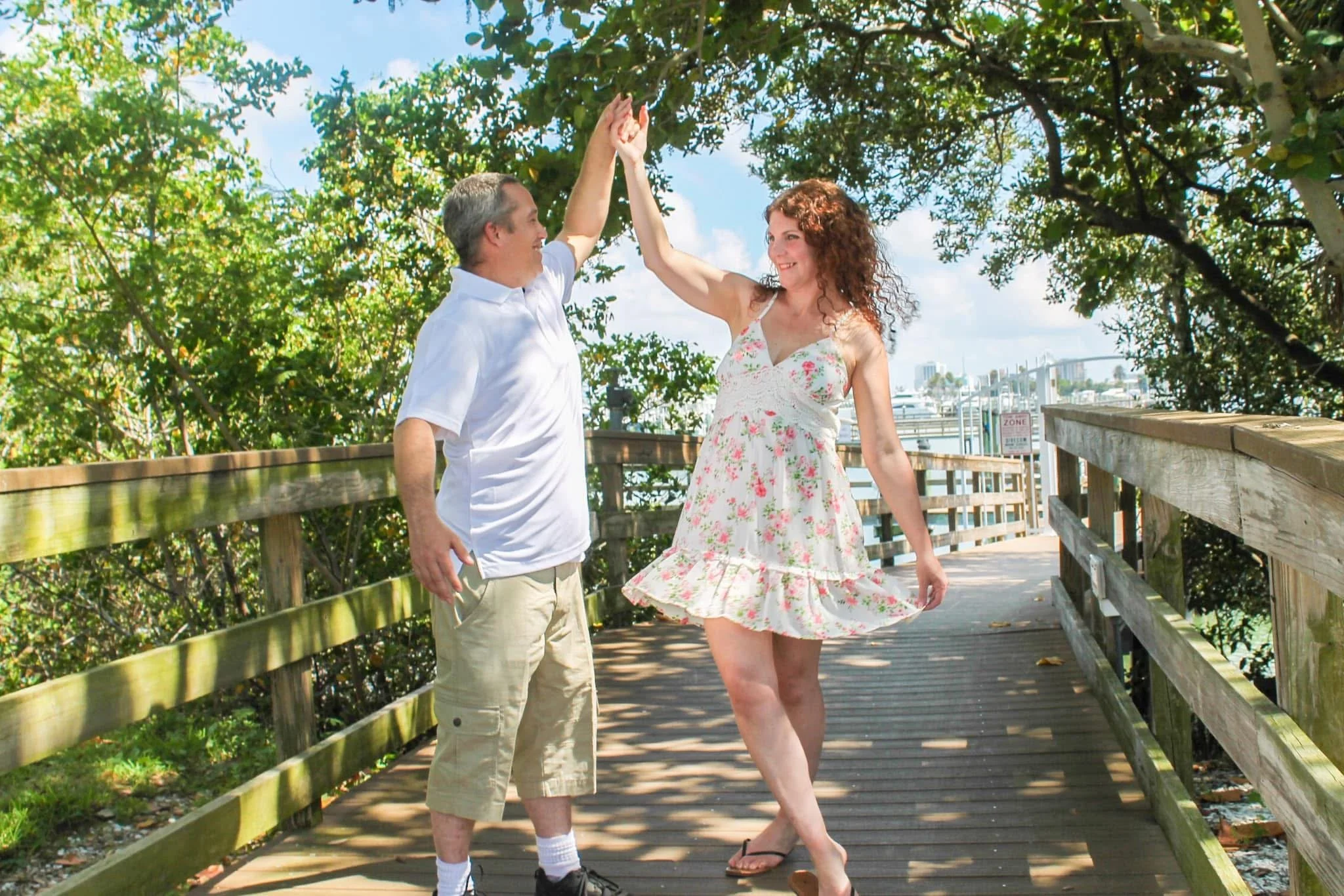 A man and woman dancing on a wooden bridge outdoors, surrounded by trees with a cityscape in the background. The man is dressed in a white shirt and khaki shorts, and the woman in a floral sundress. They are holding hands above their heads, smiling a