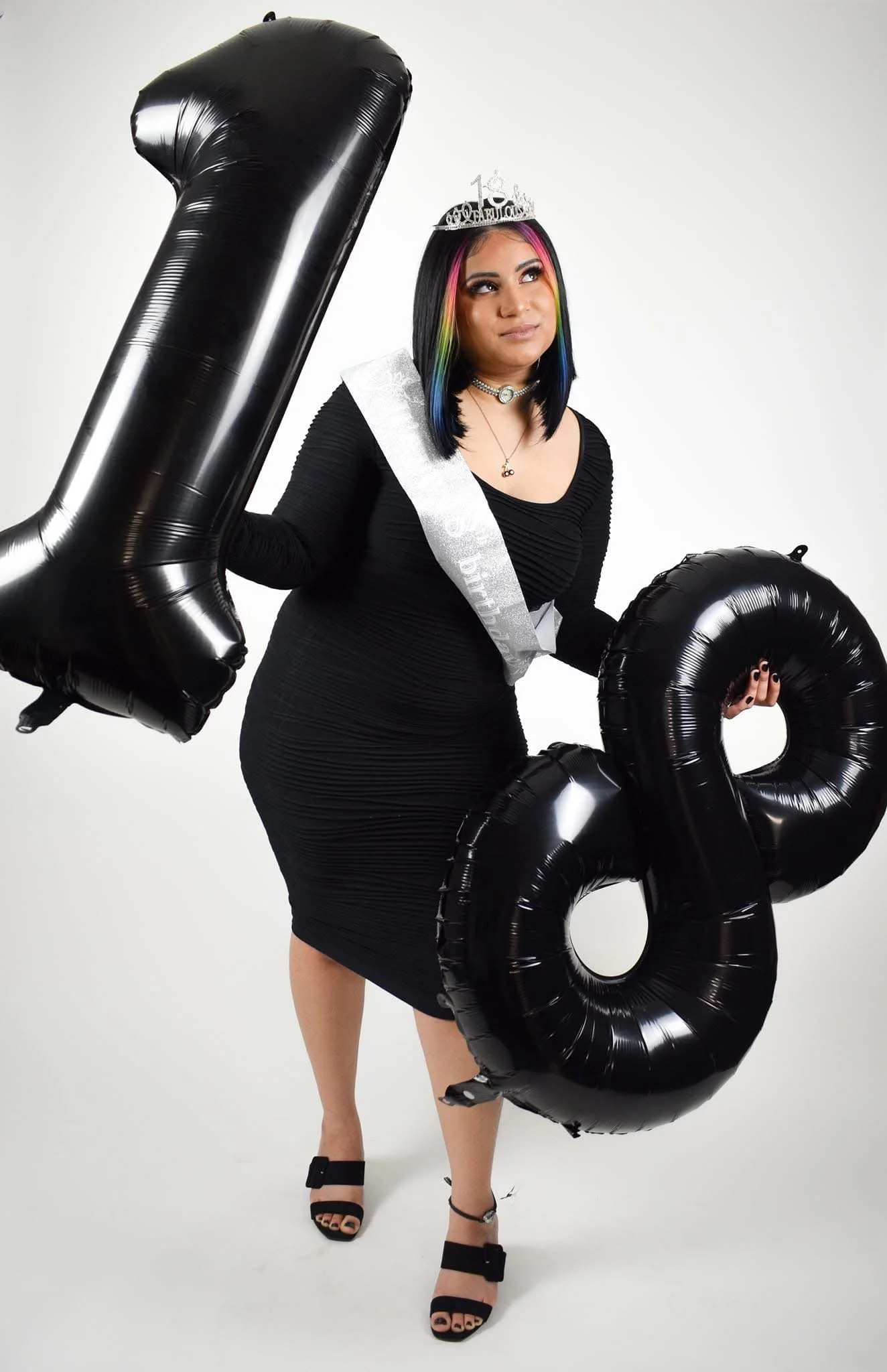 A woman celebrating her 18th birthday, wearing a black dress and birthday crown, holding large black number balloons 1 and 8.