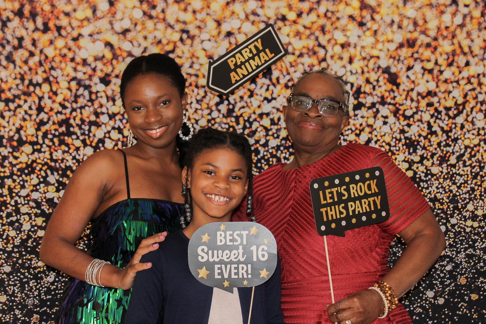 A family of three women and one girl celebrating at a party with a sparkly gold and black backdrop. They are holding signs that say "Best Sweet 16 Ever!", "Party Animal", and "Let's Rock This Party". The women are dressed in colorful attire and the g