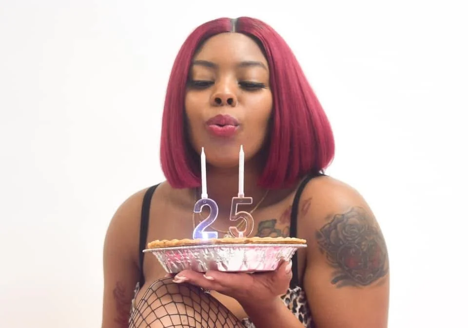 A woman with shoulder-length pink hair blowing out candles shaped as 25 on a birthday pie with candles, wearing a black top and fishnet stockings, sitting against a white background.