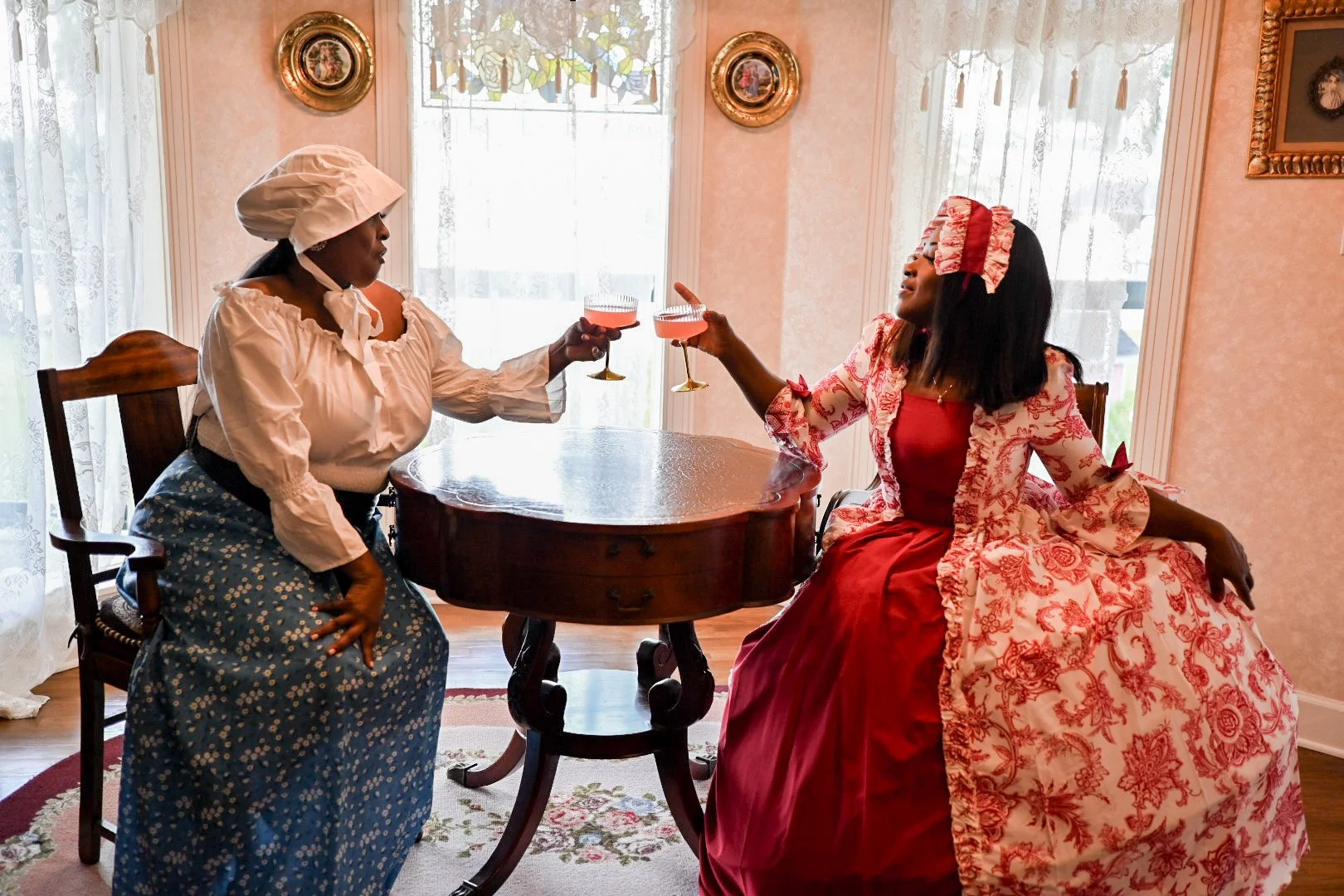Two women dressed in historical costume, toasting each other with pink drinks in a vintage-inspired room with lace curtains and framed artwork.