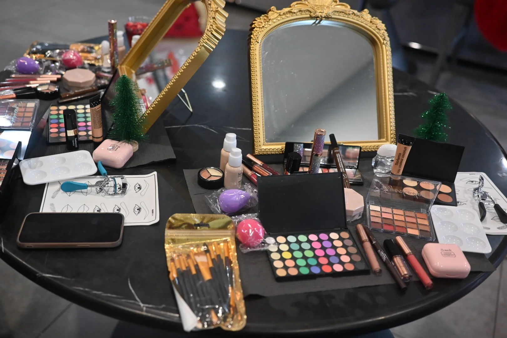 A black table covered with various makeup products including eyeshadow palettes, brushes, foundation bottles, lip glosses, a mirror with a gold ornate frame, and small decorative Christmas trees. There are also personal items like a smartphone, a pla