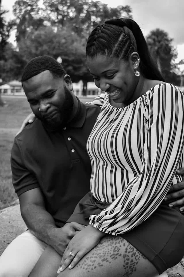 A smiling woman with braided hair and earrings is sitting outdoors with a man in a polo shirt. They are sharing a tender moment with the woman’s hand resting on her lap, which shows a ring, and the man's hand gently holding hers. Trees and a park are