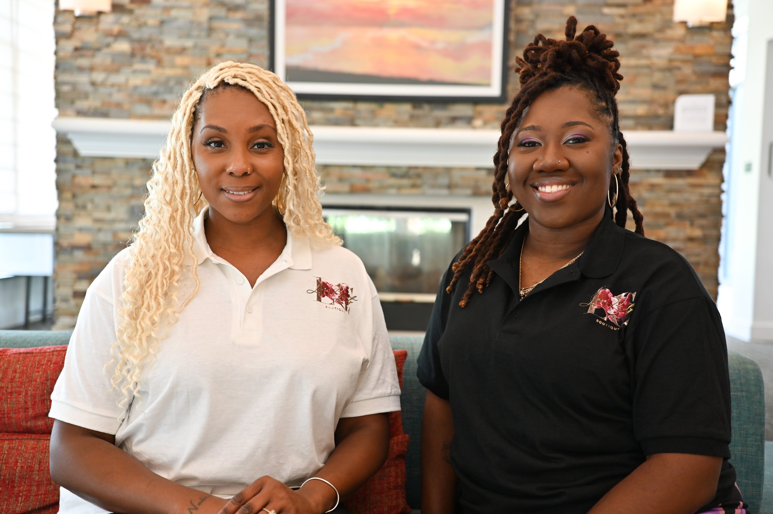 Two women sitting on a sofa in a living room, smile at the camera, with a stone fireplace and a framed painting in the background. They are wearing polo shirts with a floral logo.