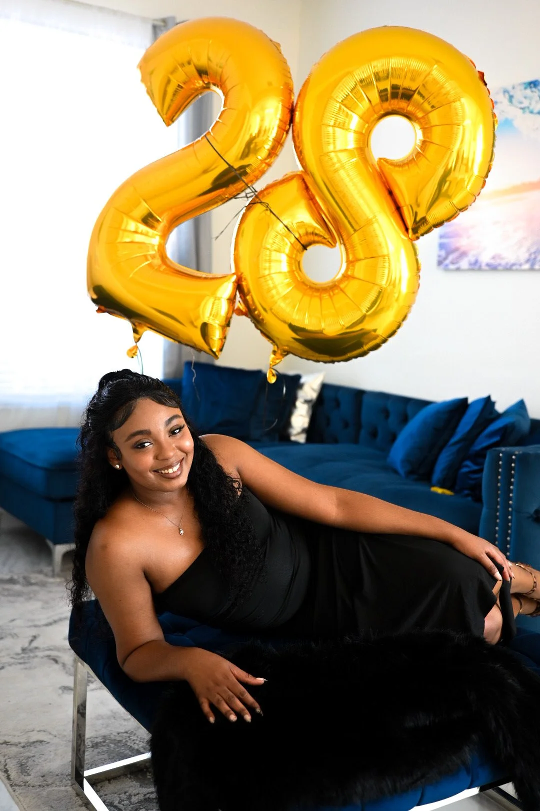 A woman in a black dress lying on a blue velvet couch with gold '2022' balloons overhead, in a living room setting.