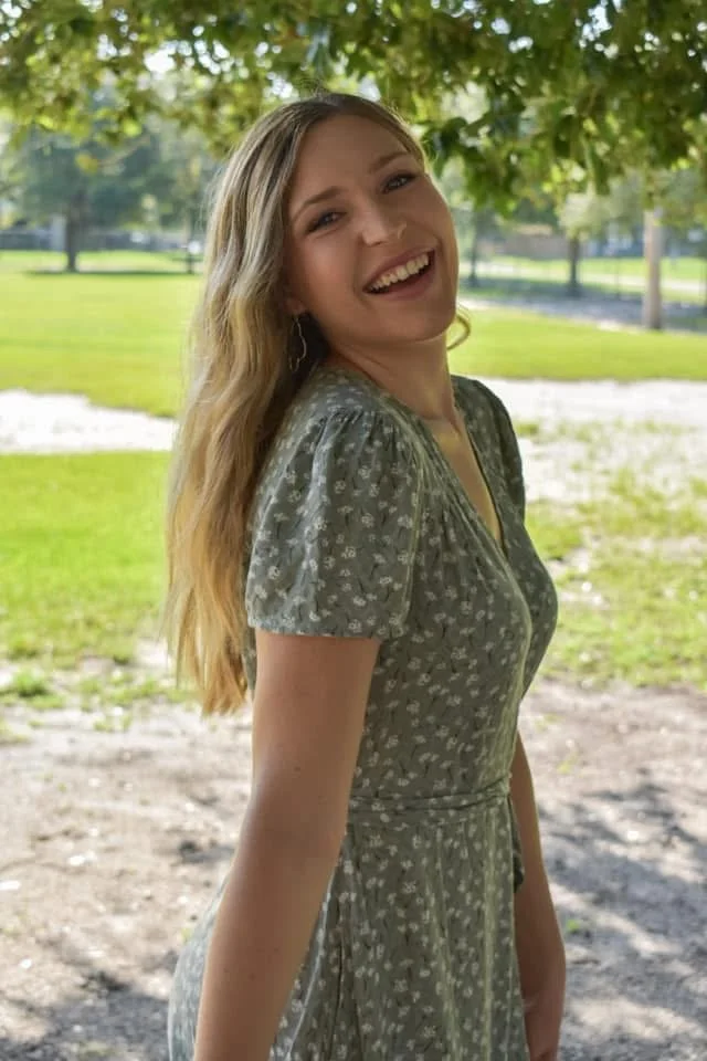 A young woman with long, wavy blonde hair standing outdoors under a tree, smiling at the camera, wearing a gray, short-sleeved dress with a small white floral pattern.