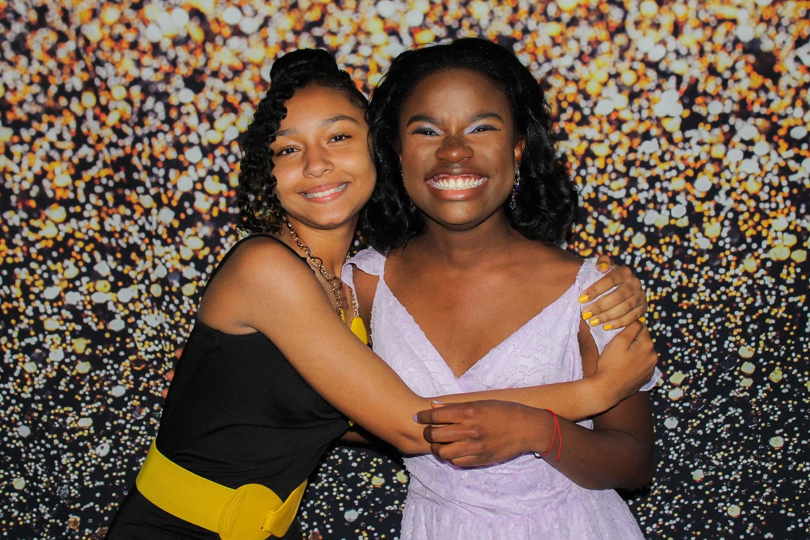 Two smiling women hugging against a sparkling gold and black background.
