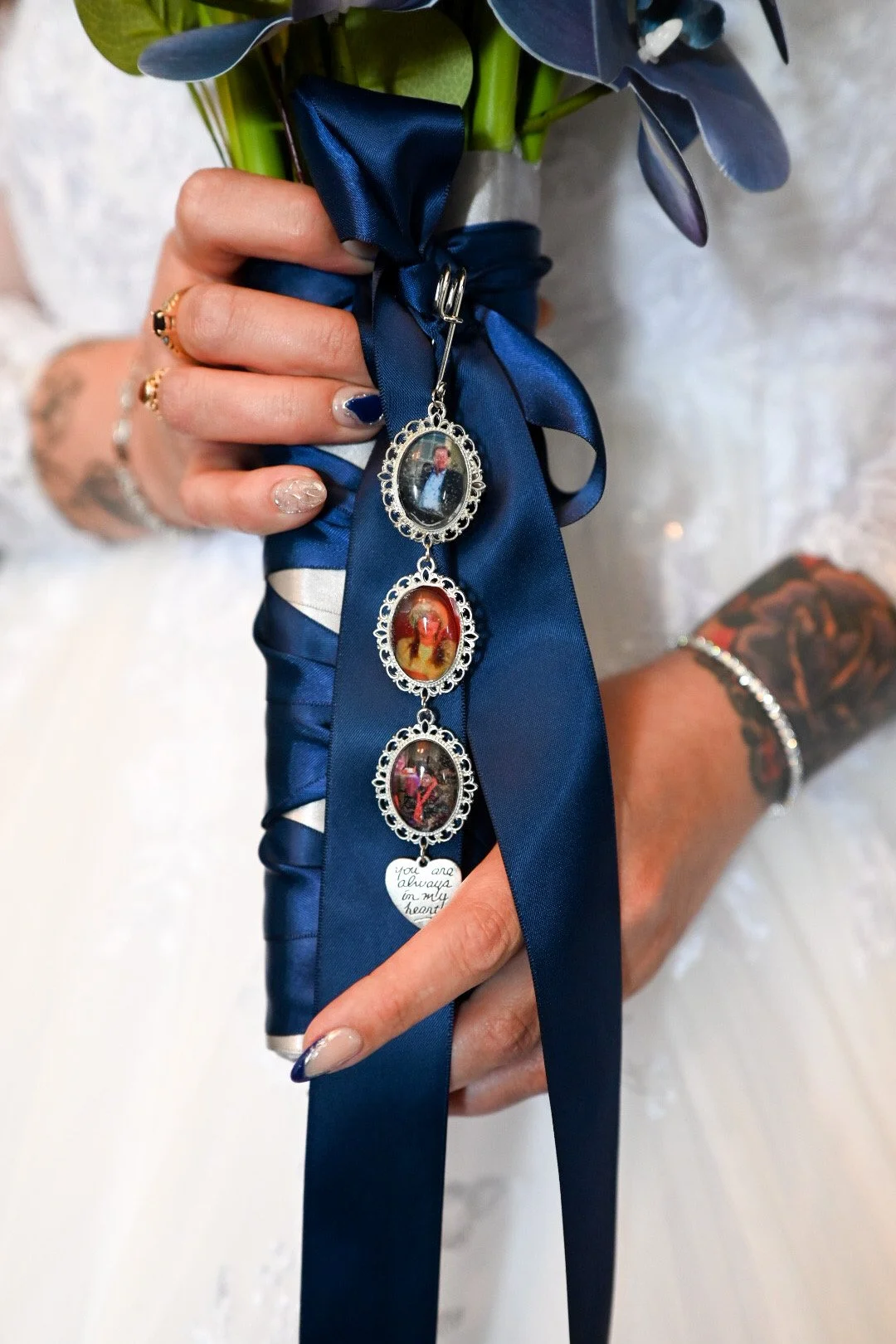 Close-up of a person's hand holding a bouquet wrapped in blue satin ribbon, with a photo charm attached. The person has painted nails, multiple rings, and a tattooed wrist, and is wearing a bracelet. The background is a white lace fabric, likely part