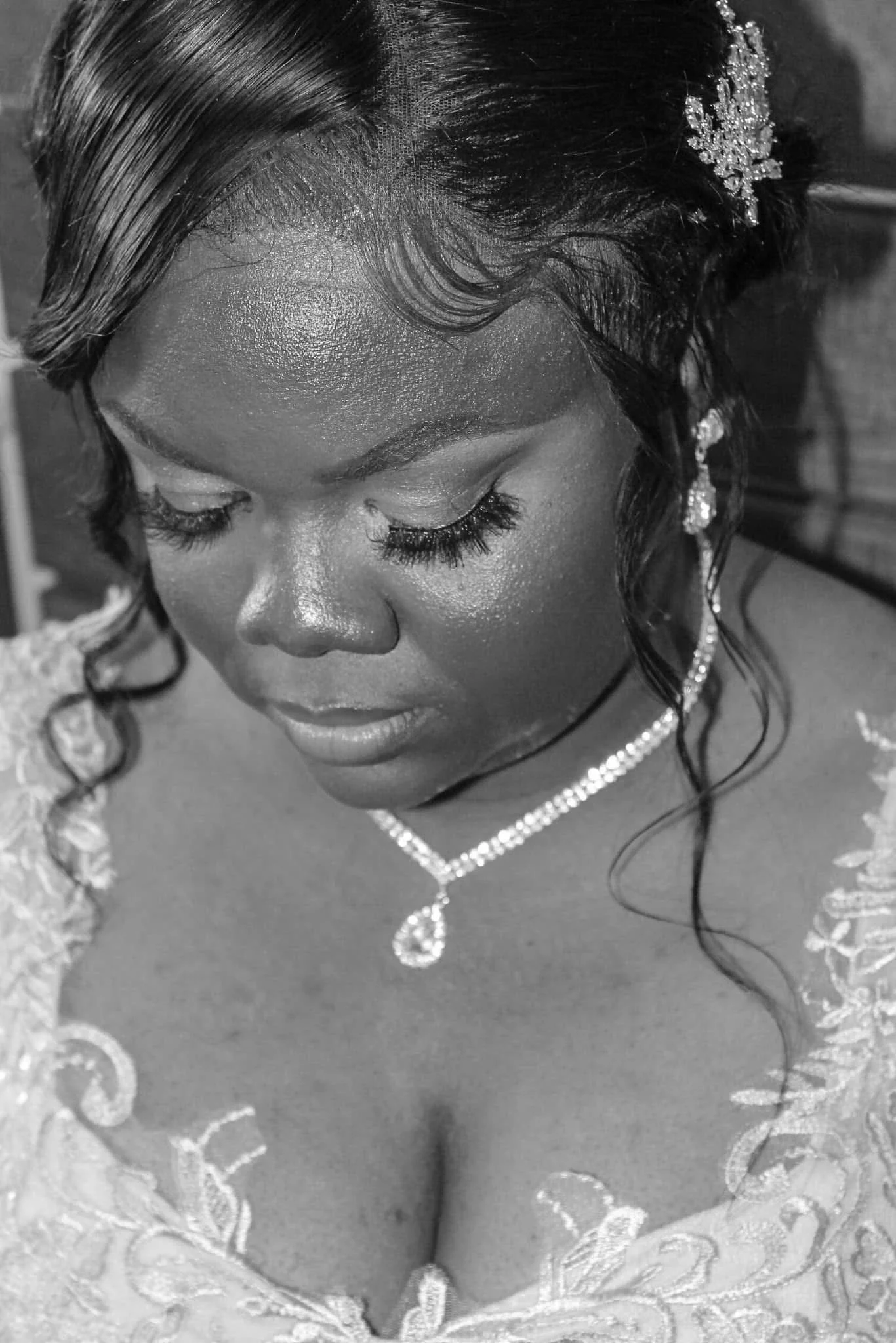 Close-up black and white photo of a woman wearing lace wedding dress, tiara, and diamond necklace, with her eyes closed and head slightly tilted downward.