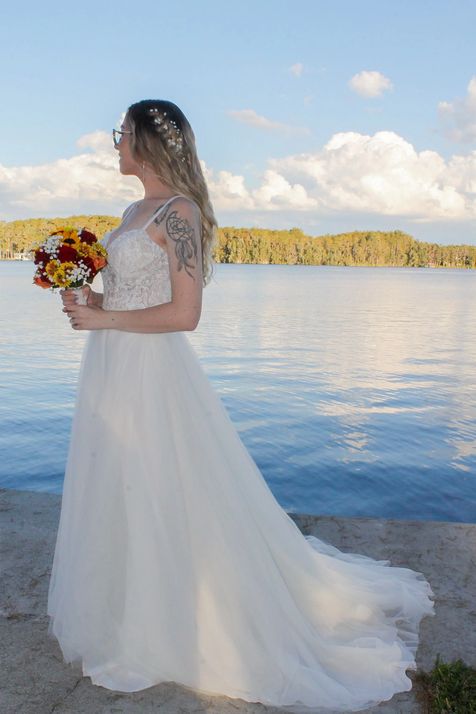 A woman in a wedding dress holding a bouquet of flowers standing by a lake during daytime.