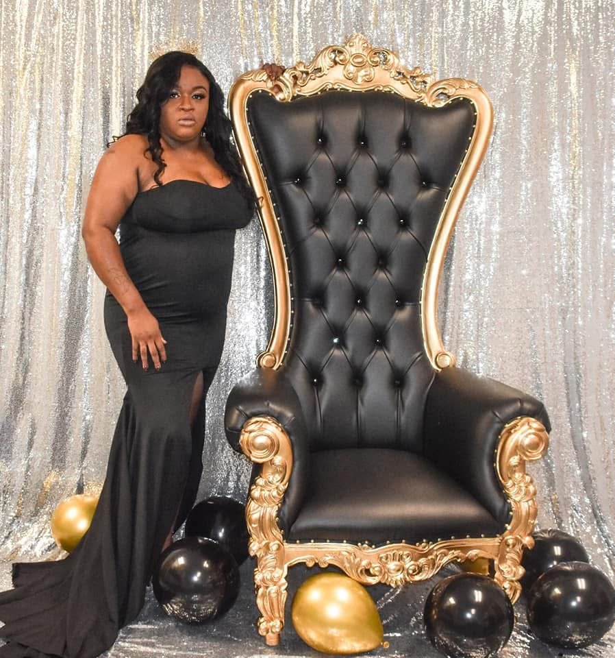 A woman in a long black gown standing next to a large, ornate black and gold throne with black and gold balloons on the floor, in front of a shimmering silver backdrop.