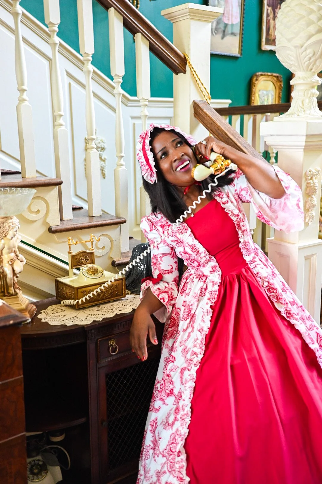 A woman dressed in a 19th-century style pink gown and matching bonnet, talking on a vintage rotary phone in a decorated house interior.