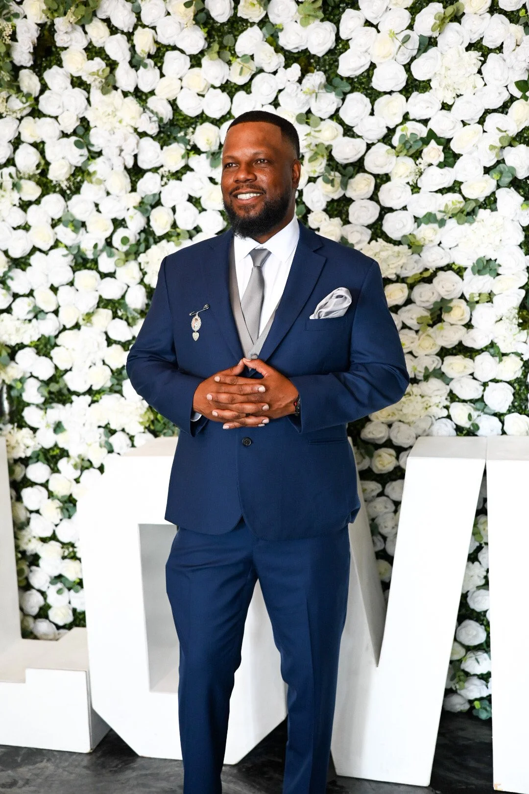 A man dressed in a navy blue suit, white shirt, silver tie, and pocket square, standing in front of a white flower wall with large white letters partially visible, smiling confidently.