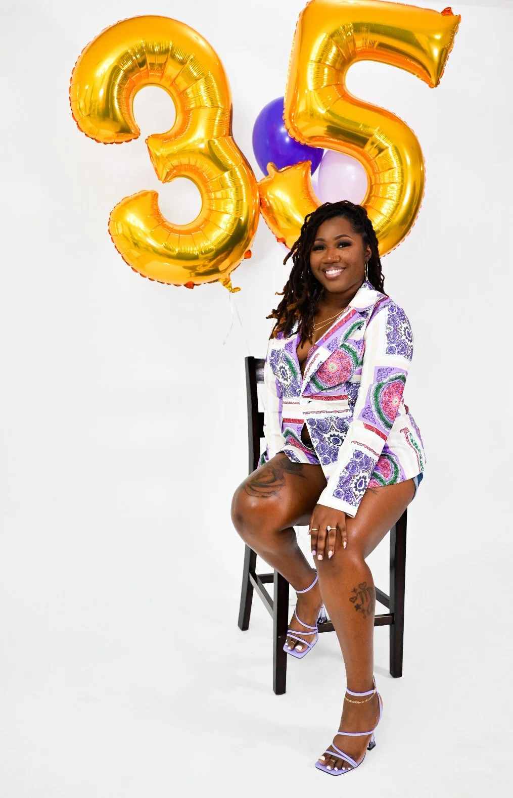 A woman sitting on a chair with a smile, wearing a colorful patterned blazer and shorts. She has shoulder-length curly hair and visible tattoos on her legs. Behind her are gold balloons shaped as the number 35.