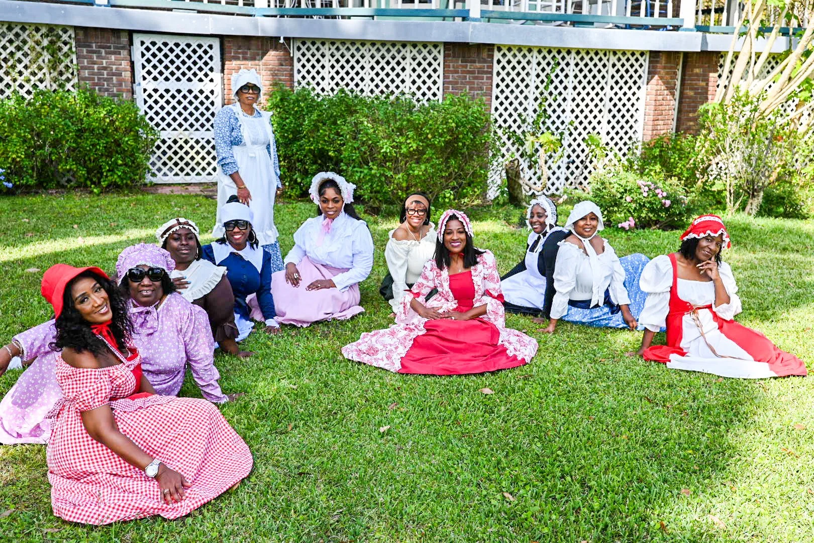 A group of women dressed in historical costumes sitting and kneeling on grass in a garden, with a brick building and white lattice in the background.