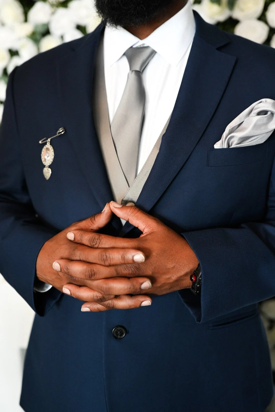 A man in a navy blue suit, white shirt, and gray tie with a pocket square, clasping his hands together.