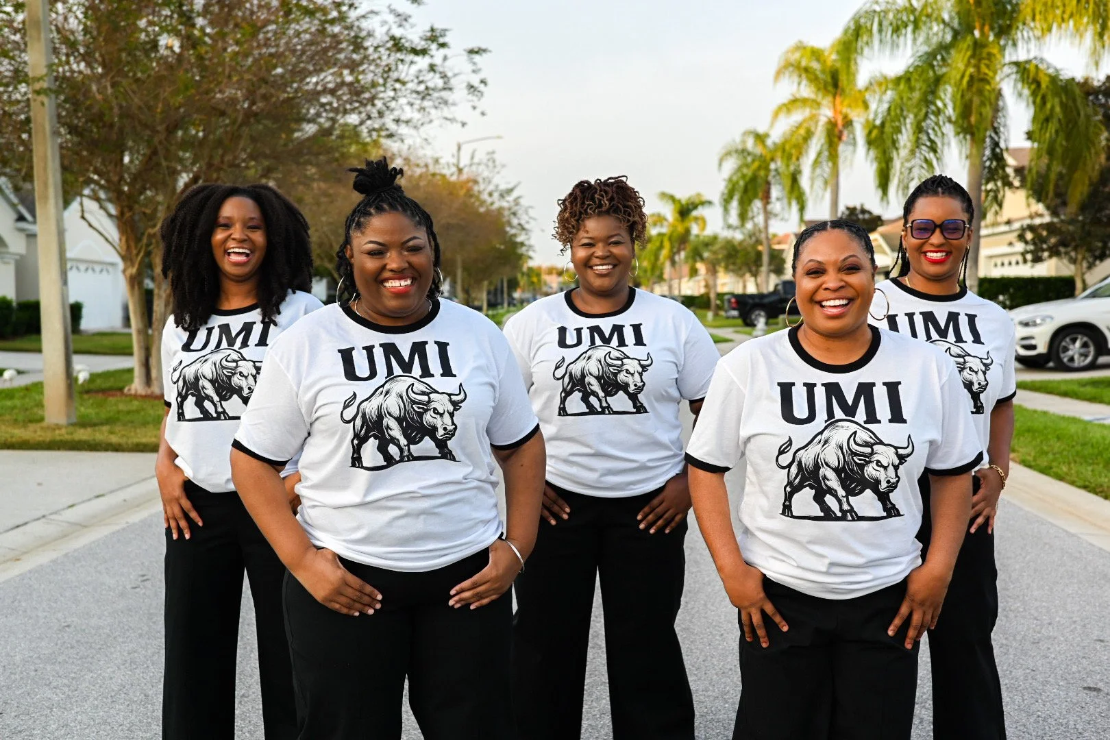 Group of five women standing outdoors on a residential street, wearing matching white t-shirts with 'UMI' and a bull graphic, smiling and posing for the photo.