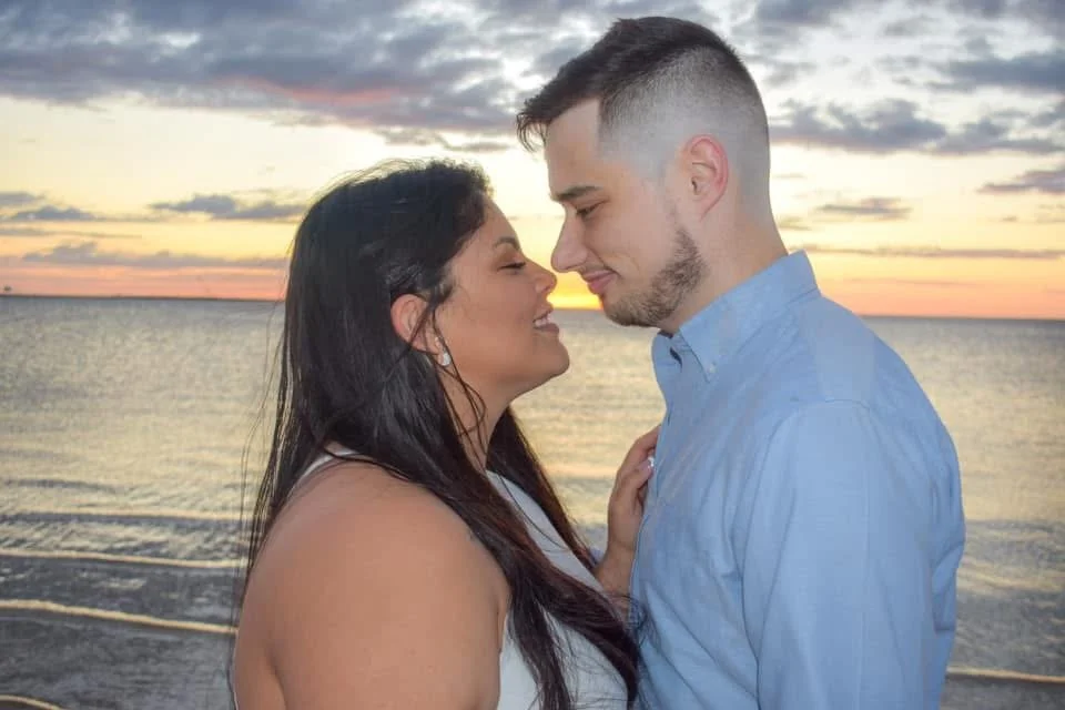 A couple standing close to each other on a beach at sunset, with their faces touching and smiling.