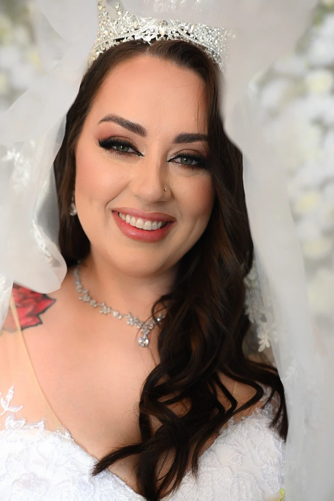 A woman with dark wavy hair wearing a tiara, white wedding dress, and jewelry, smiling for a portrait.
