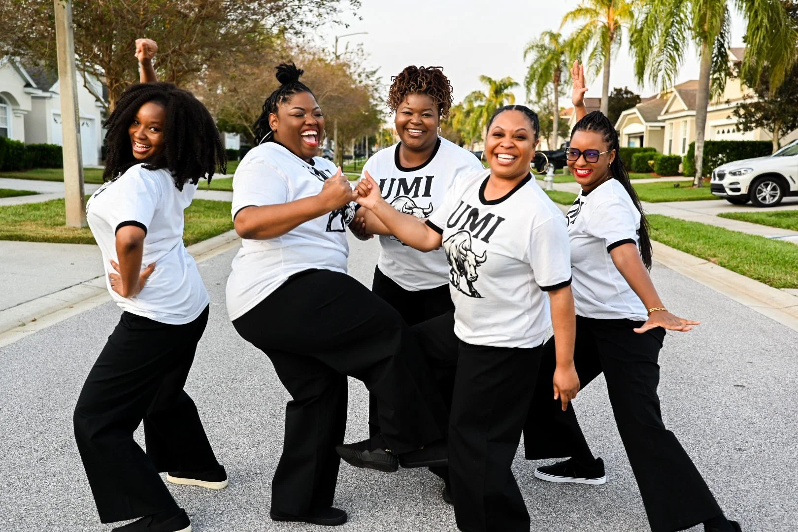 Five women wearing matching white shirts with the UMI logo, standing on a suburban street, smiling and making playful poses.