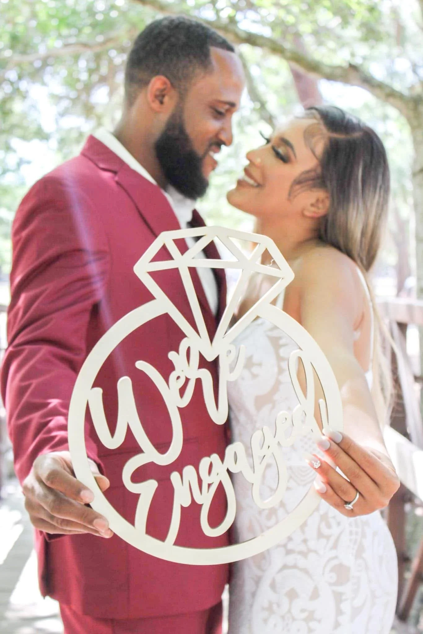 A bride and groom holding a circular sign with a diamond and the words 'We Said Yes' cut out, standing outdoors with trees in the background, smiling at each other.