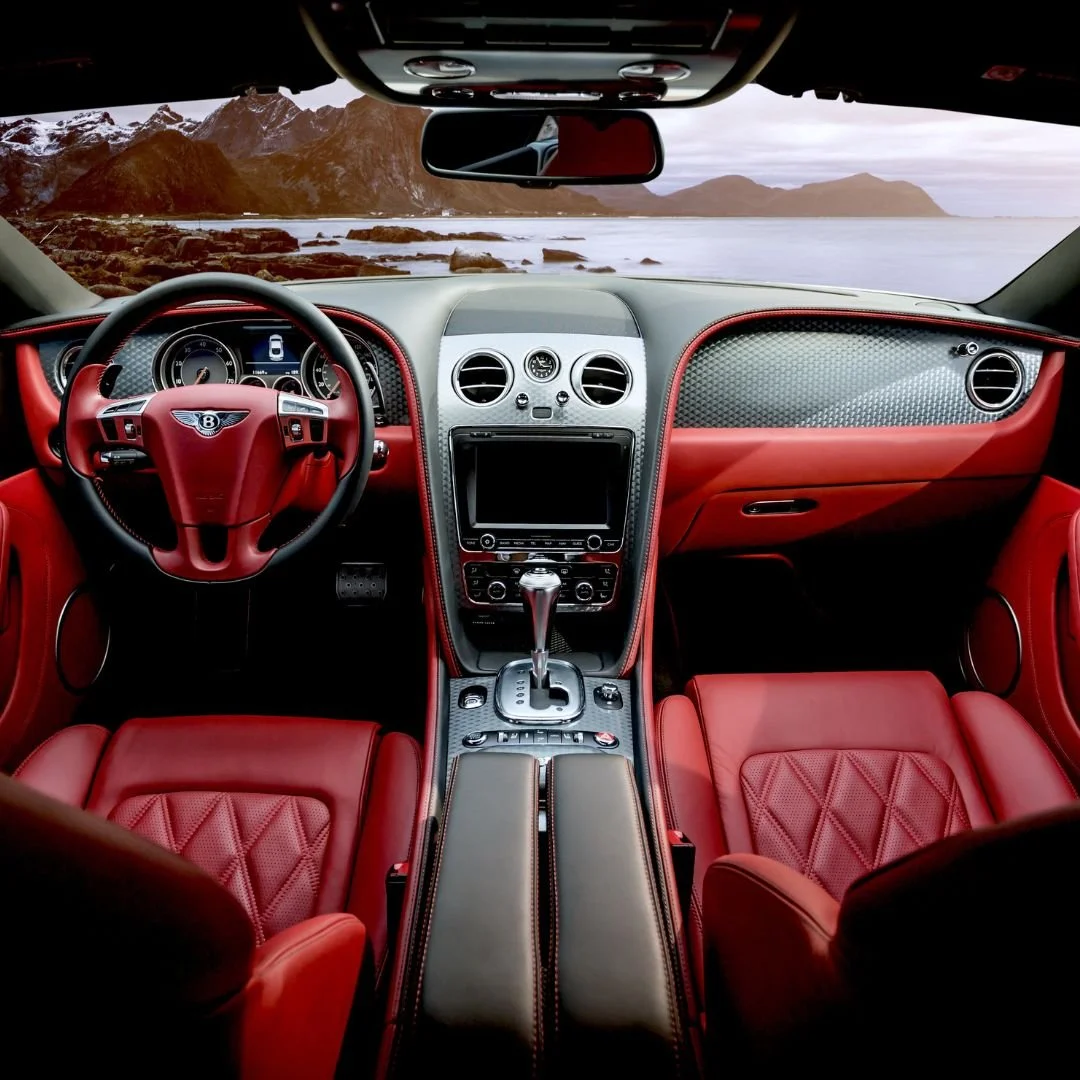 Interior of a luxury car with red leather seats, a carbon fiber dashboard, and a scenic ocean view with mountains visible through the windshield.