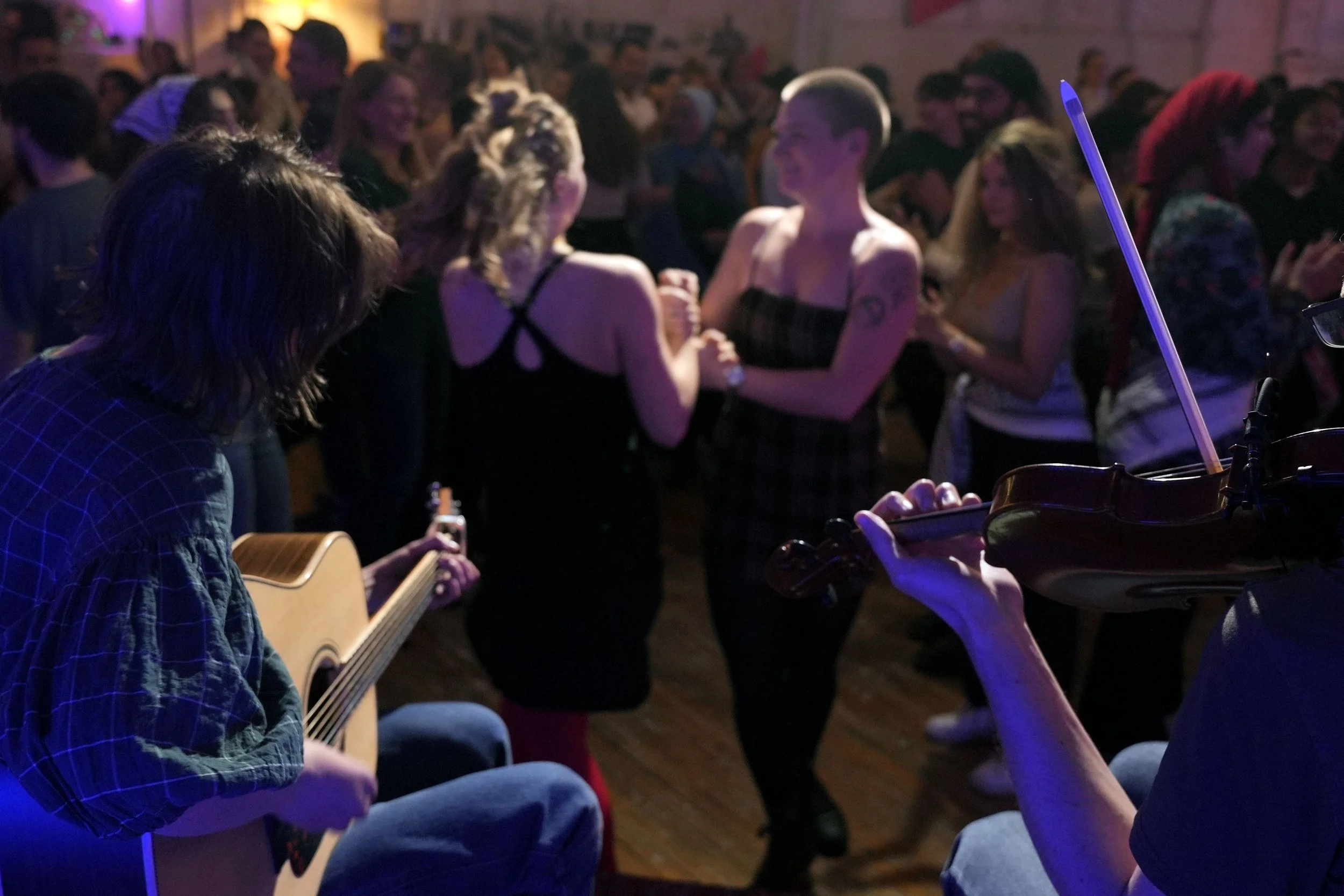 People dancing and enjoying live music in a dimly lit indoor venue, with a musician playing the acoustic guitar and another person playing the violin.