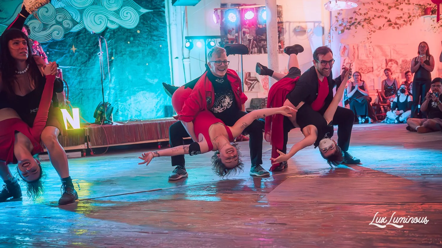 People performing acro yoga on a wooden dance floor, with some dressed in red and black outfits, in a colorful indoor space with audience watching, colorful lighting, and decorations on the walls.