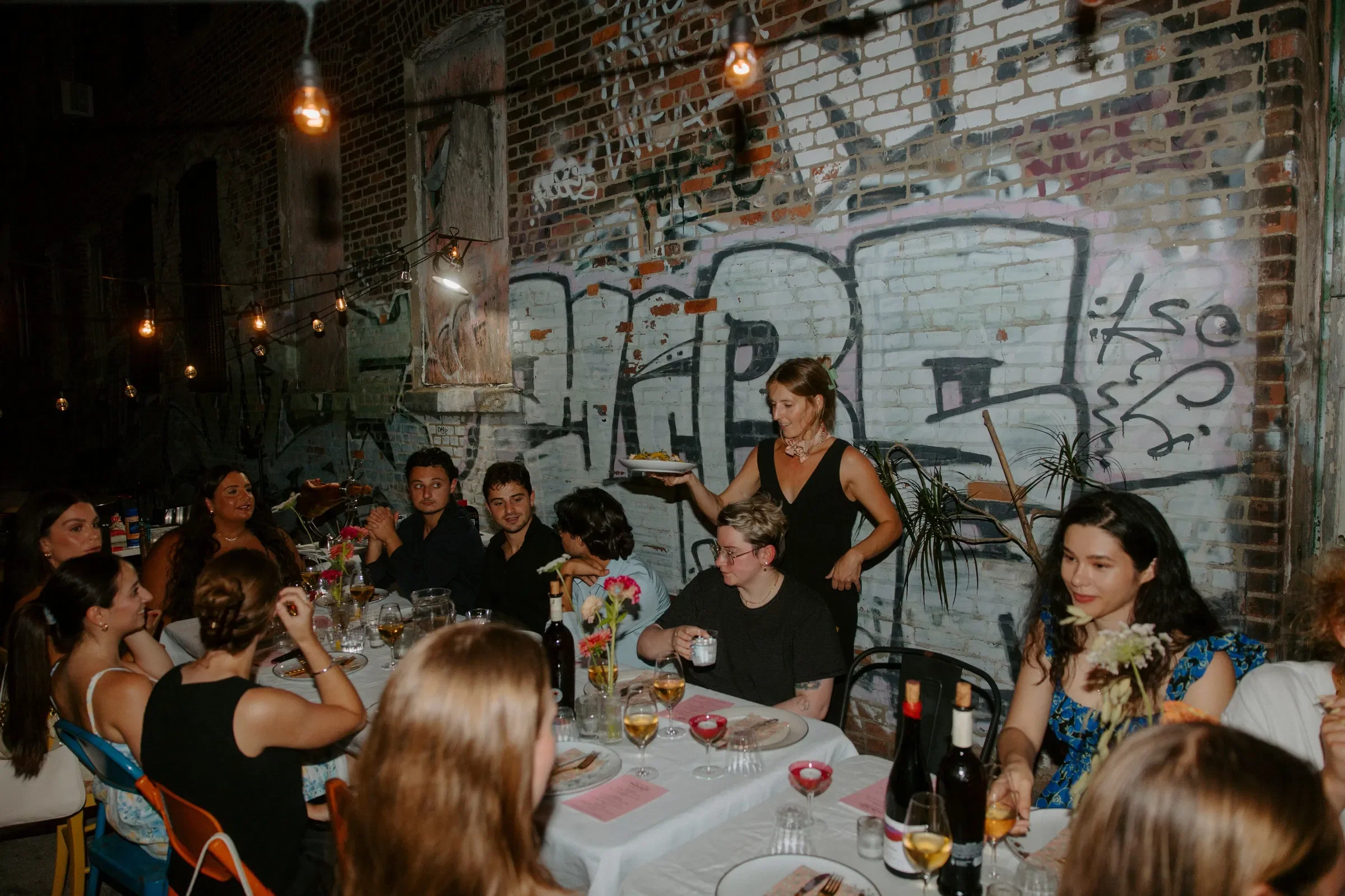 Woman in black dress serving food to a group of people seated at a dinner table in an industrial-style restaurant with graffiti on brick walls.