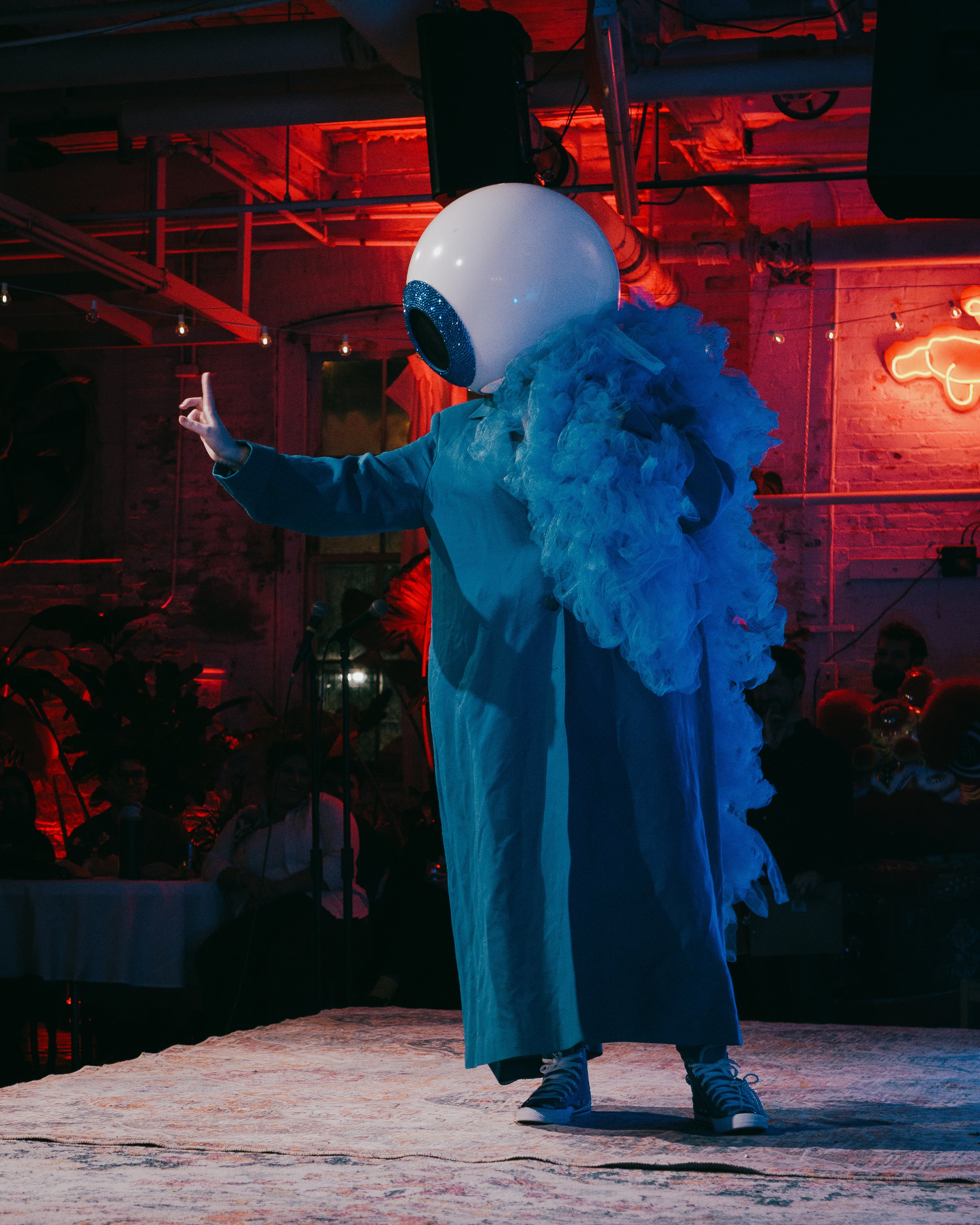 Person wearing a large eyeball mask, ruffled blue shirt, oversized overcoat, and sneakers, standing on a stage with a dark, neon-lit background.