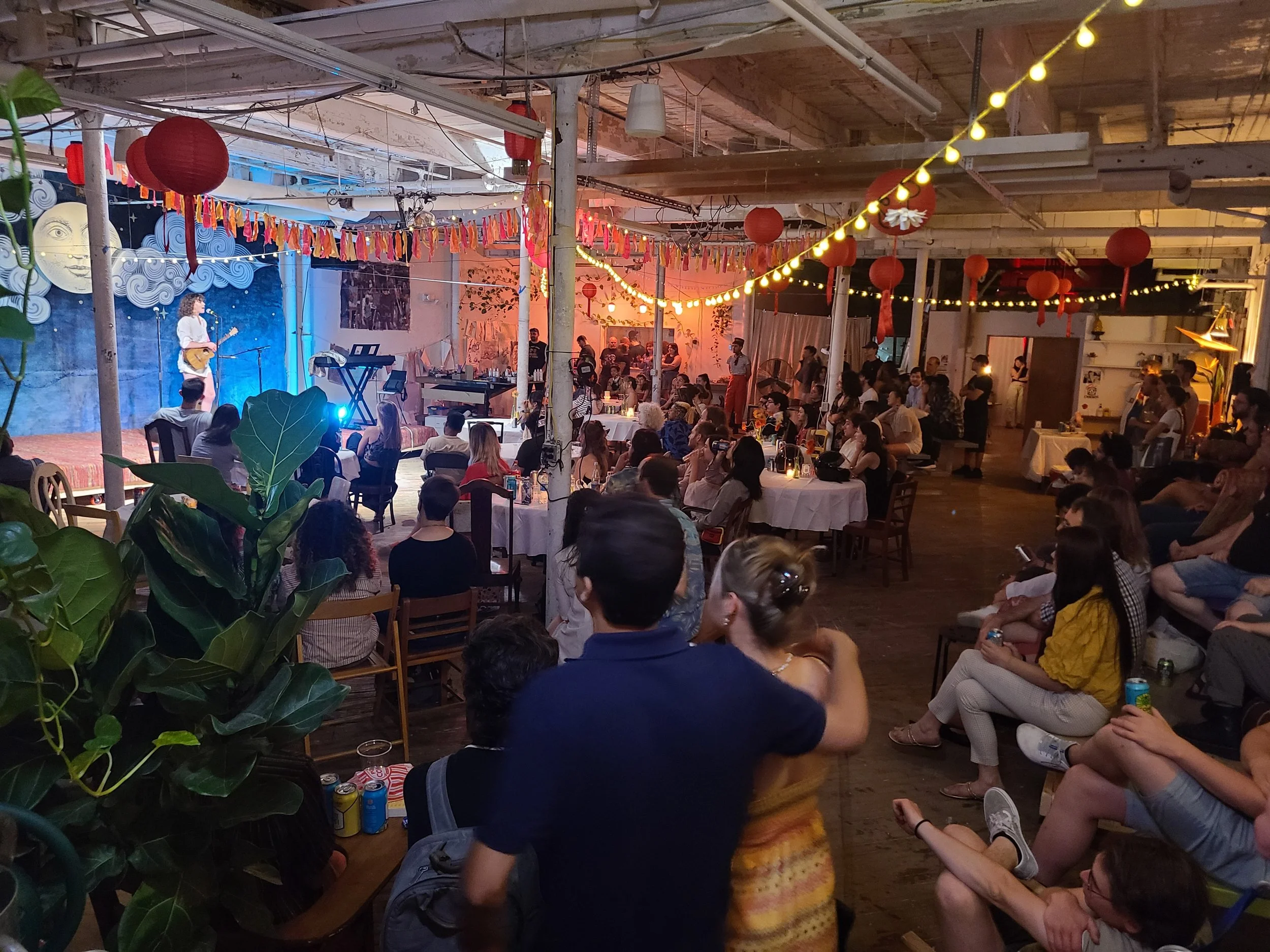 A lively indoor event with a stage for live music, decorated with colorful banners, paper lanterns, and string lights. Audience members sit at tables and on chairs, watching performers on stage in a rustic, warehouse-style space with wood beams and a painted mural backdrop.