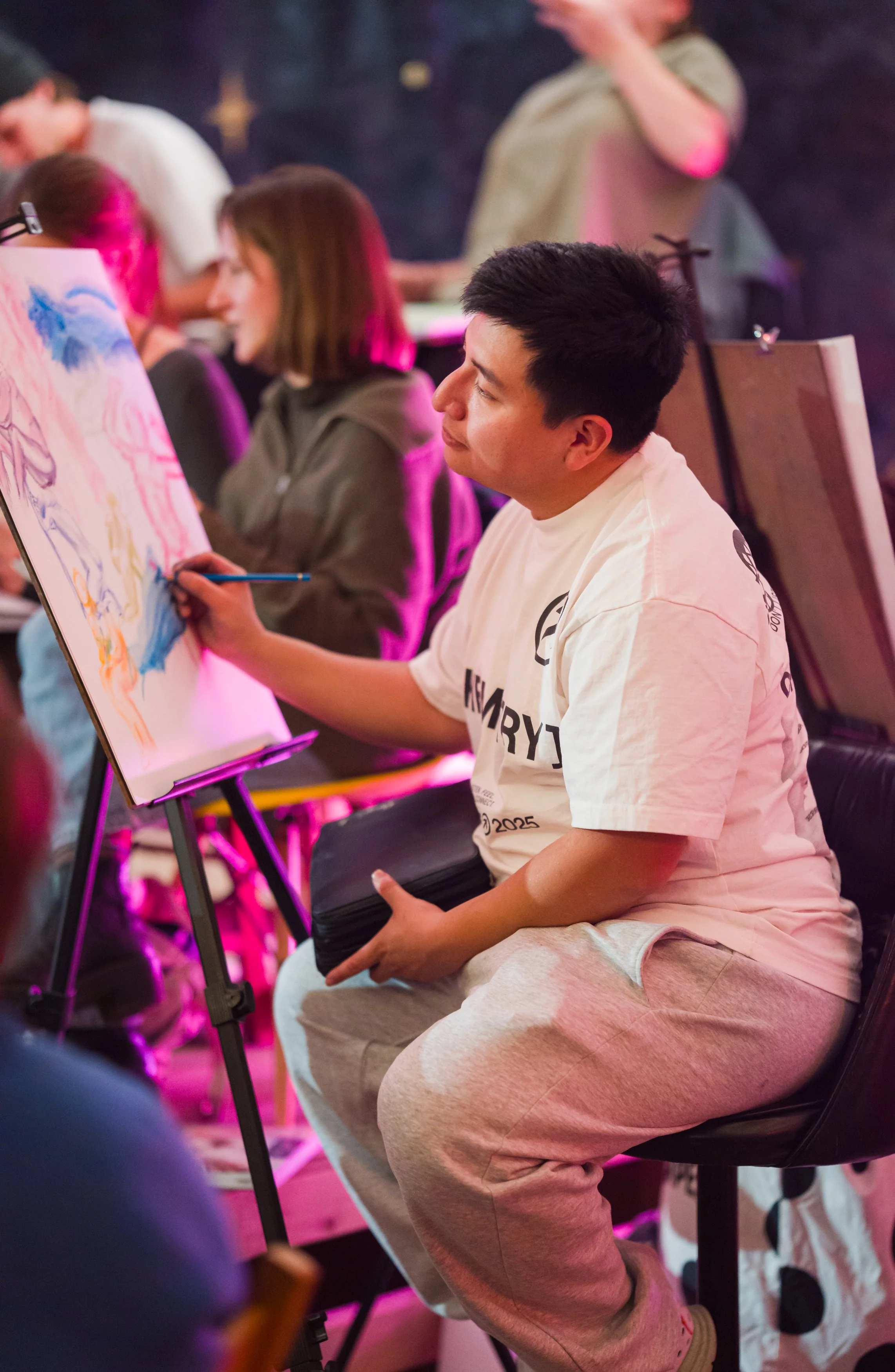 A man painting on a canvas at an art event, surrounded by other people, with pink and purple lighting.