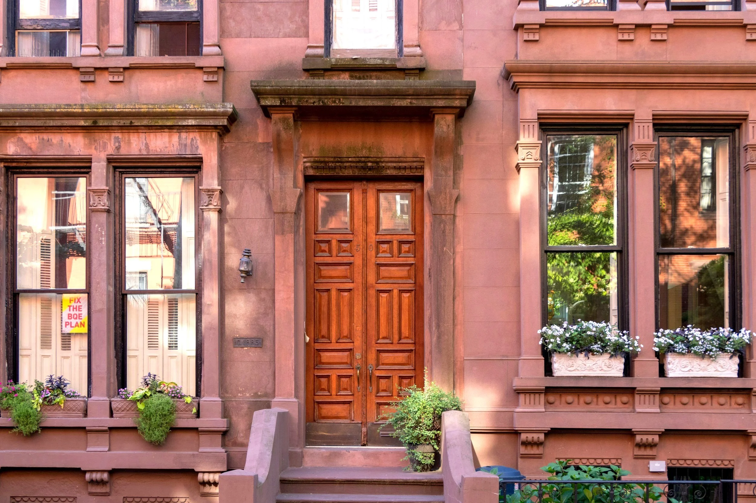 Brownstone townhouse with ornate carved window trim, wooden front door, potted plants, and flower boxes in a city neighborhood.
