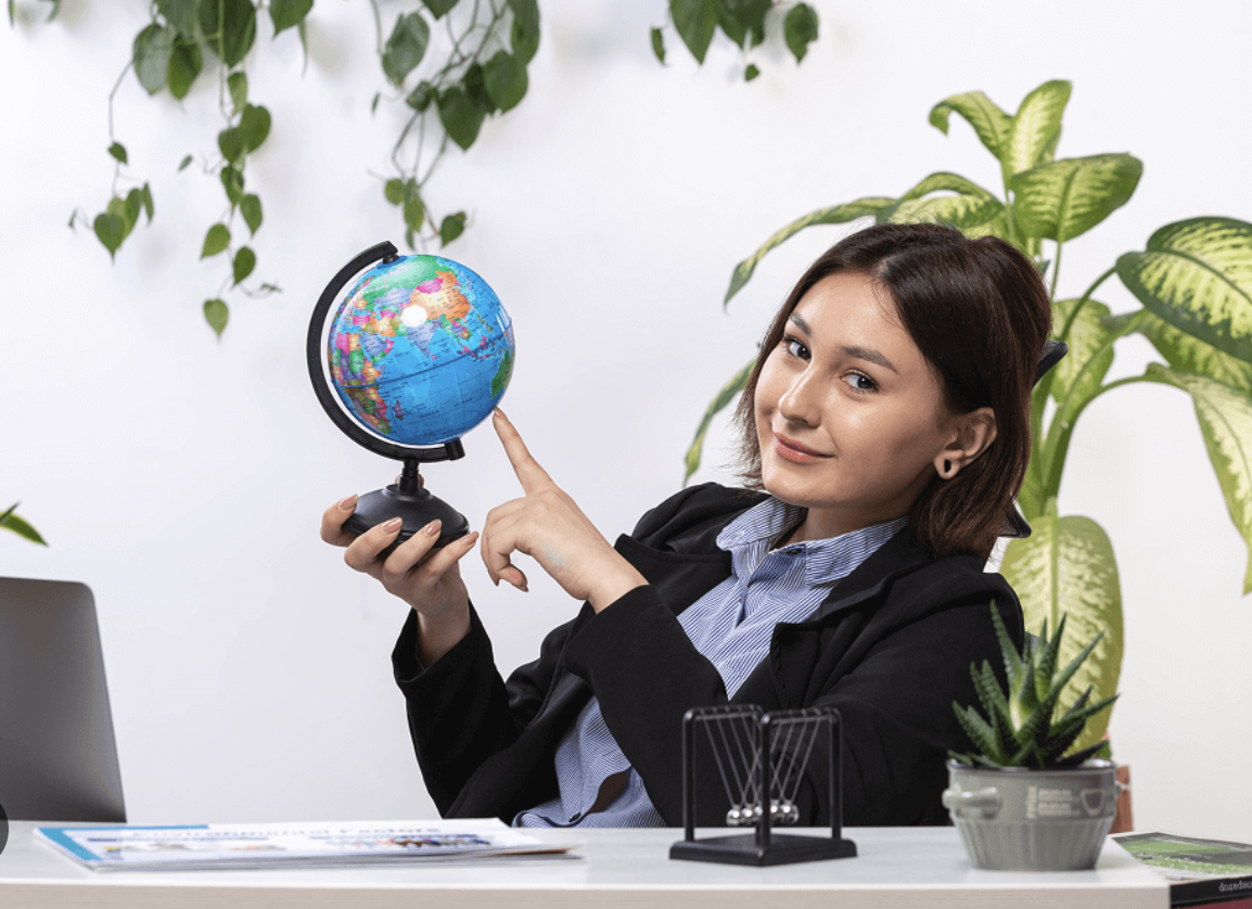 A woman in business attire sitting at a desk with a globe in her hand, pointing to it, surrounded by green plants.